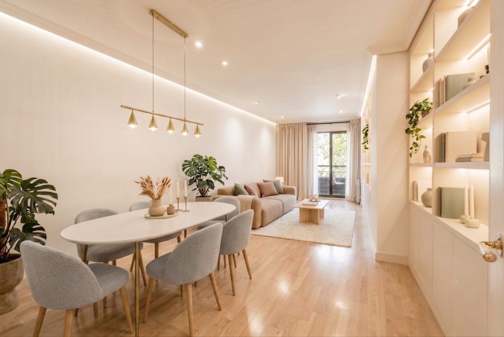 Salón comedor moderno reformado con mesa ovalada blanca, sillas de bouclé grises, lámpara colgante dorada y estantería blanca con iluminación LED integrada.