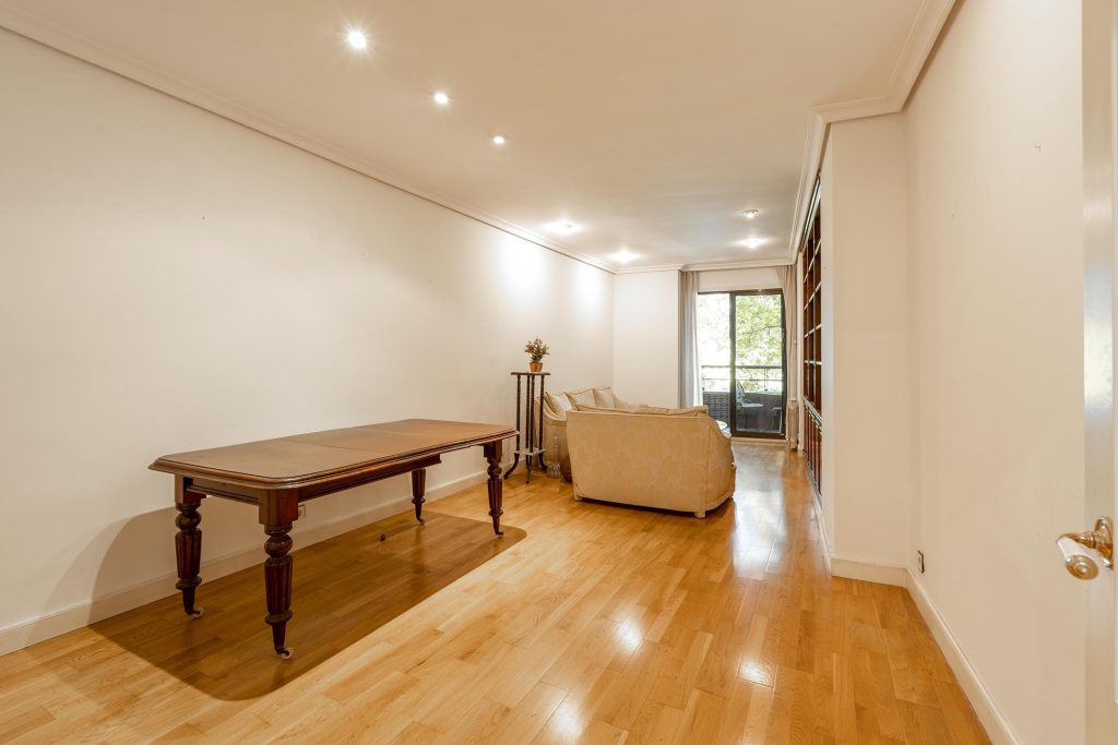 Estado original del salón comedor antes de la reforma, con mesa de madera oscura, sofá beige antiguo e iluminación básica mediante focos empotrados.